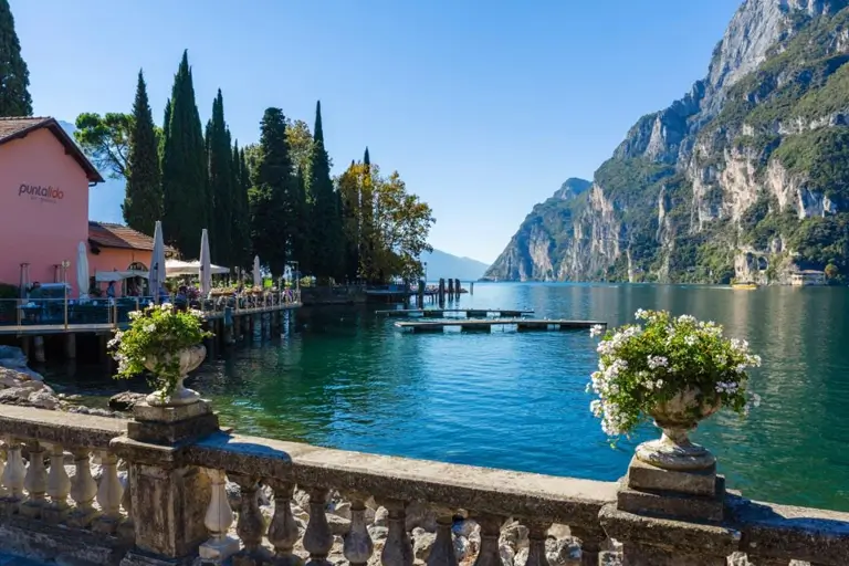 Scenic lakeside view with cypress trees, colorful buildings, and steep mountains reflecting in the calm water of Lake Garda.