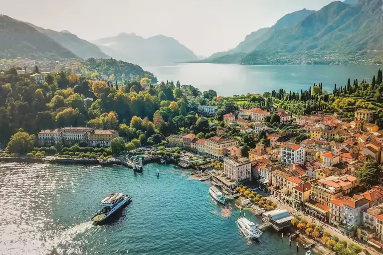 A picturesque view of a lakeside town surrounded by mountains, with vibrant buildings, boats, and lush greenery along the water's edge.