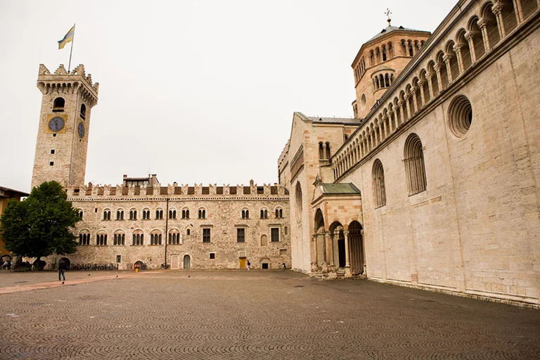 Wide stone plaza with historic cathedral building, arched colonnades and medieval clock tower in a quiet Italian city square